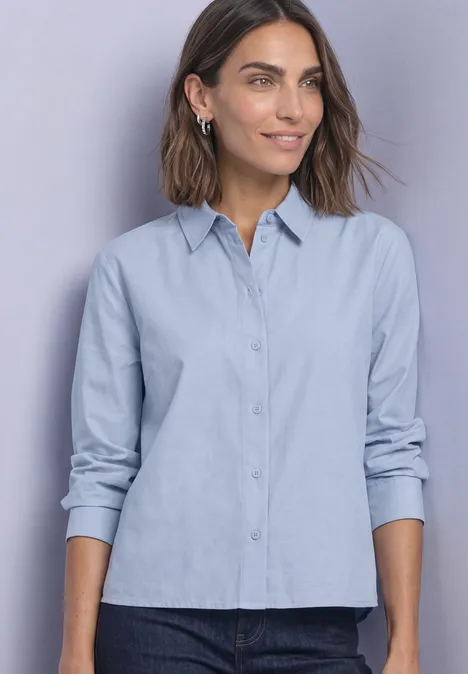 Femme portant une chemise bleu clair à manches longues et un jean foncé sur fond neutre.