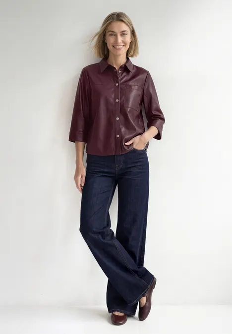 3/4-Arm Hemdbluse im Leder-Look rustic rouge Frau in burgunderrotem Hemd aus Leder und weiten Jeans steht vor weißem Hintergrund, lächelnd und lässig.
