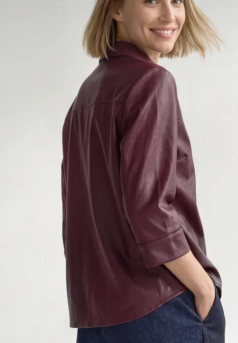 3/4-Arm Hemdbluse im Leder-Look rustic rouge Frau in weinroter Lederbluse und Jeans, Hände in Taschen, lächelt über die Schulter vor weißem Hintergrund.