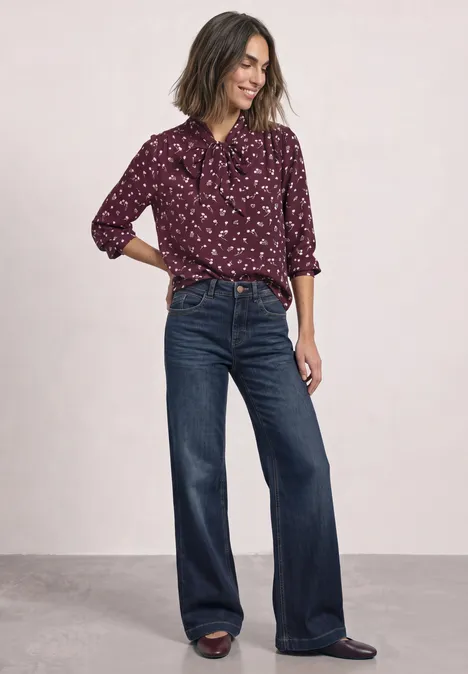 Eine Frau trägt eine bordeauxrote gemusterte Bluse und weite Jeanshose.