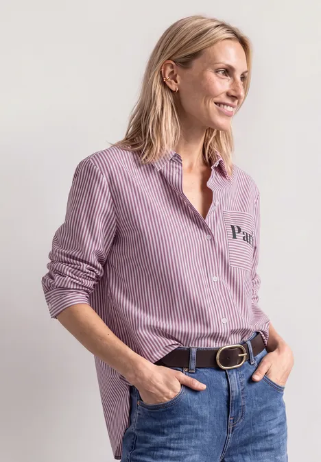 Frau in gestreiftem rosafarbenem Hemd und Jeans lehnt lässig, Hände in den Hosentaschen, Blick zur Seite.