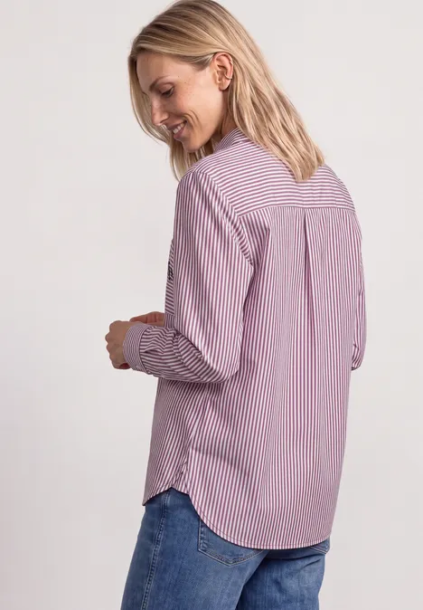 Eine blonde Frau trägt eine rosa-weiß gestreifte Bluse und Jeans, steht seitlich und lächelt.