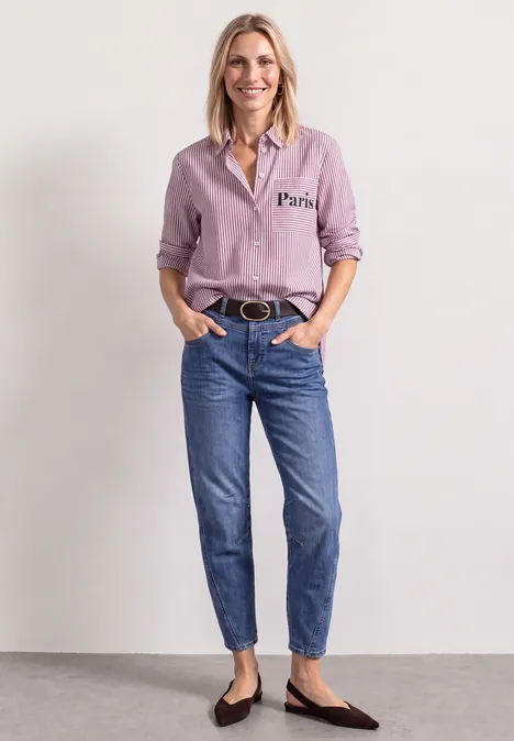 Frau in rosa Bluse, blauen Jeans und schwarzen Ballerinas vor neutralem Hintergrund.