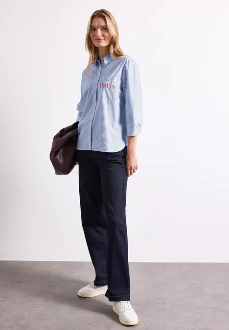 Langarm Hemdbluse mit Wording Detail northern blue Eine Frau trägt eine hellblaue gestreifte Bluse, dunkelblaue Hose und weiße Sneakers, in der Hand eine Jacke.
