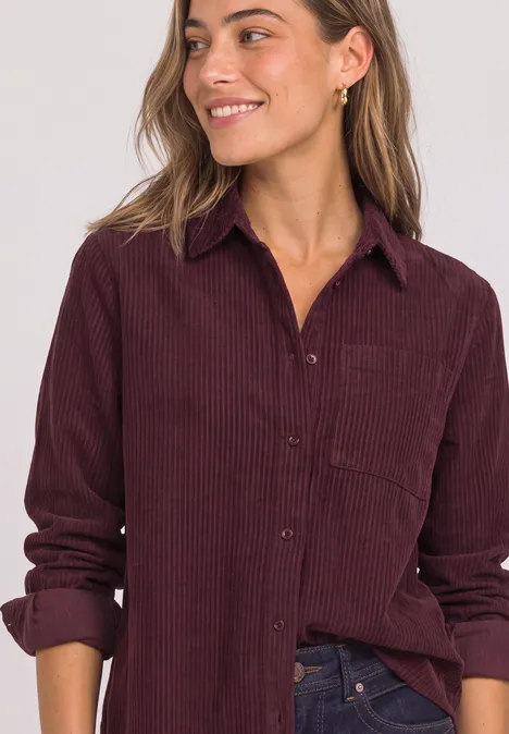 Une femme souriante porte une chemise en velours côtelé bordeaux et un jean foncé.