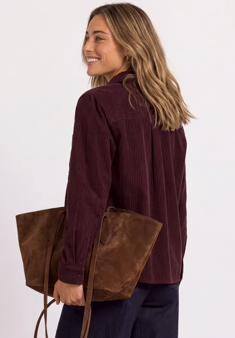 Femme portant une chemise en velours côtelé bordeaux, souriante, tenant un grand sac en daim marron.
