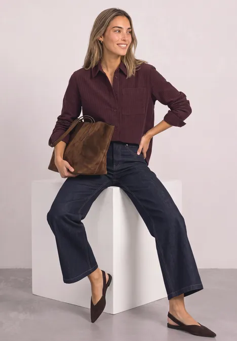 Femme portant une chemise en velours côtelé bordeaux, jean large et mules, tenant un sac en nubuck marron devant un fond clair.