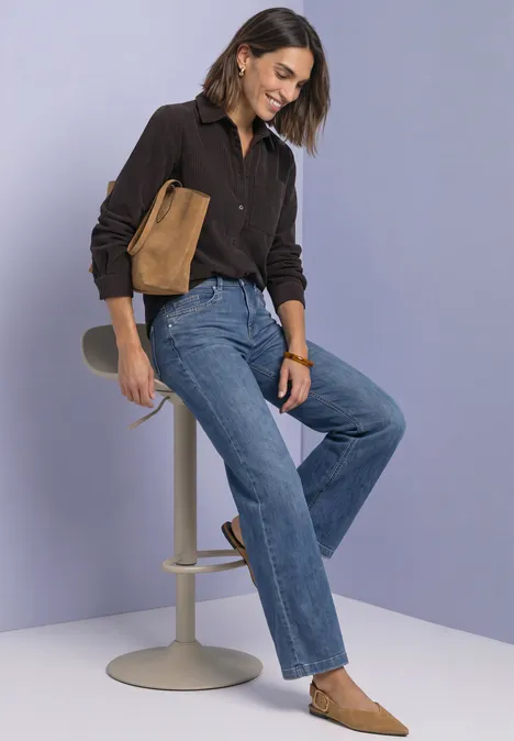 Femme en jean et chemise brune avec sac assorti, assise sur un tabouret, fond clair, style décontracté.