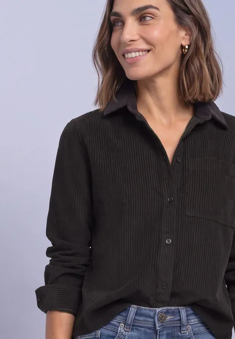 Femme souriante aux cheveux bruns mi-longs porte une chemise foncée à col avec manches retroussées.