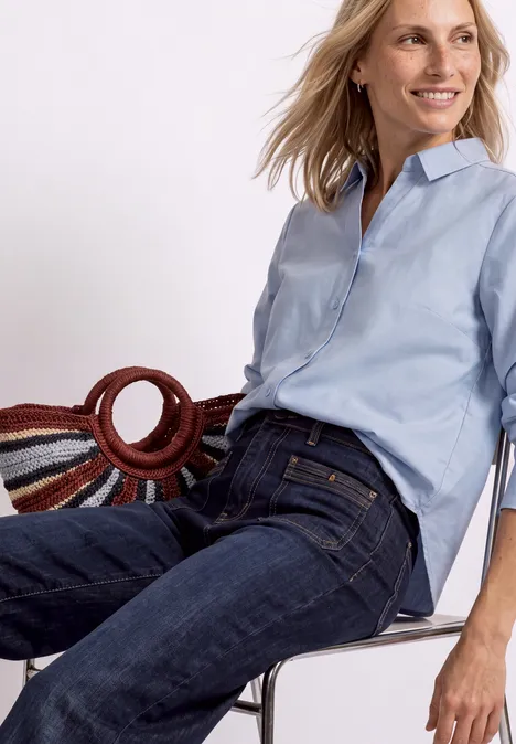 Femme souriante en chemise bleu clair et jean foncé assise sur une chaise.