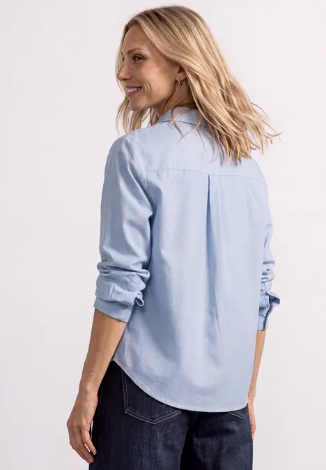 Femme vue de dos portant une chemise bleu clair, tournant légèrement la tête.