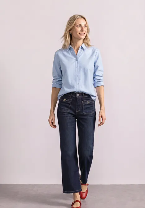 Femme en chemise bleu clair, jean foncé et sandales rouges, posant devant un fond neutre.