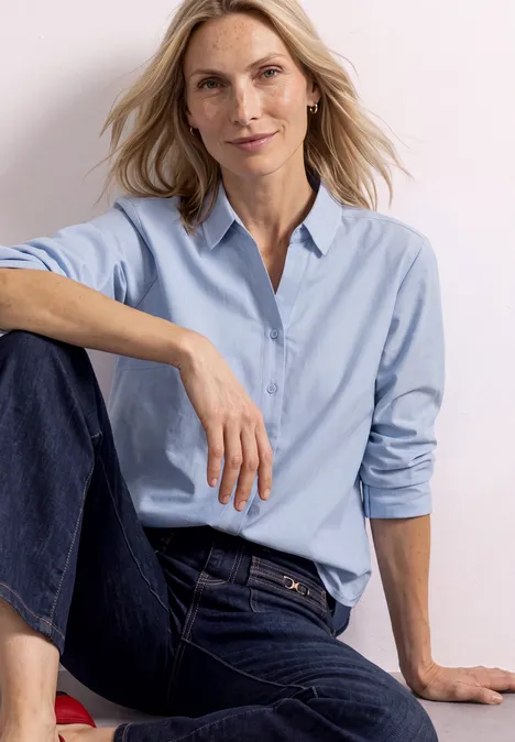 Femme blonde en jean et chemise bleu clair, assise et détendue, sourit à l'appareil photo.