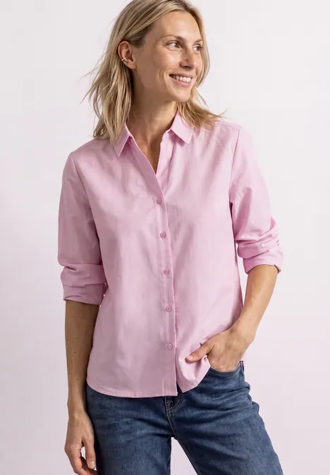 Une femme souriante portant une chemise rose et un jean sur fond clair.