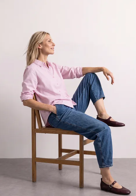 Une femme en chemise rose et jean est assise détendue sur une chaise en bois, souriante de profil.