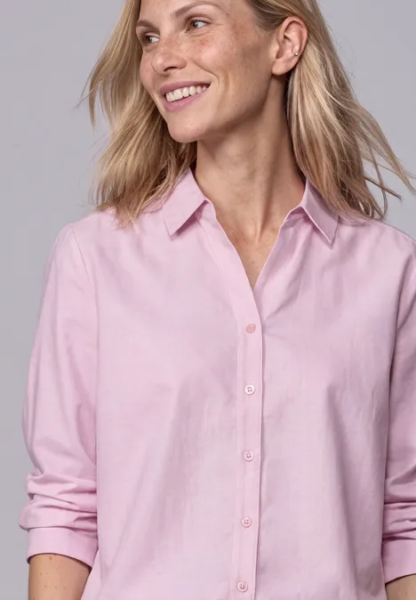 Une femme aux cheveux blonds porte une chemise rose clair et sourit.