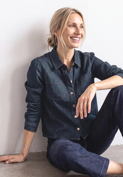 Femme souriante assise sur le sol devant un mur, vêtue d’une chemise et d’un jean en denim.