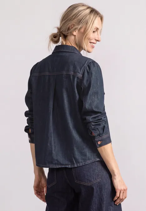 Femme vue de dos, chignon blond, chemise en jean foncé et jean, sourire latéral.