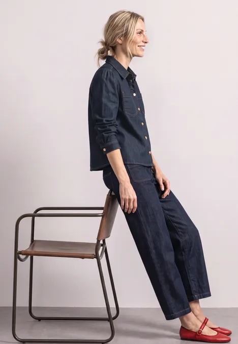 Femme appuyée sur une chaise, porte un ensemble en jean foncé et des ballerines rouges.