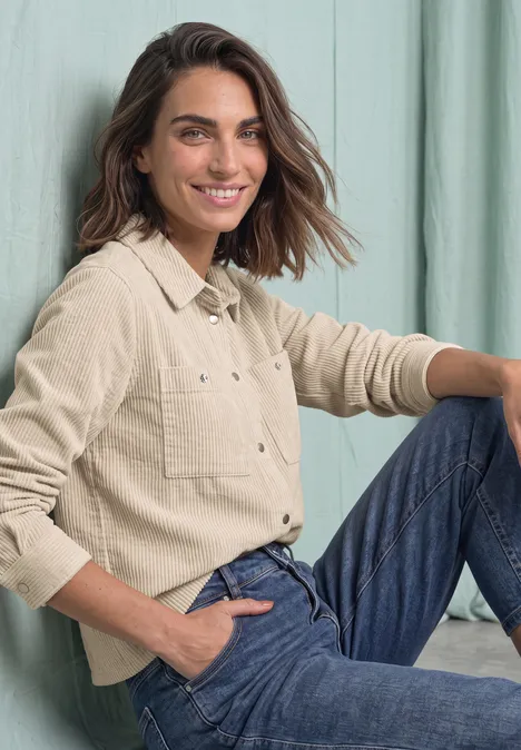 Eine Frau mit braunen Haaren sitzt in Jeans und beigem Hemd lächelnd vor einem hellen Hintergrund.