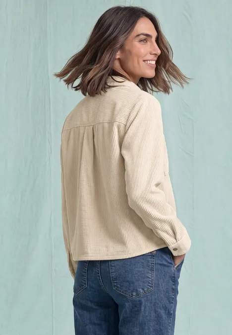 Eine lächelnde Frau mit braunen schulterlangen Haaren, trägt eine beige Jacke und Jeans, seitlich vor hellem Hintergrund.