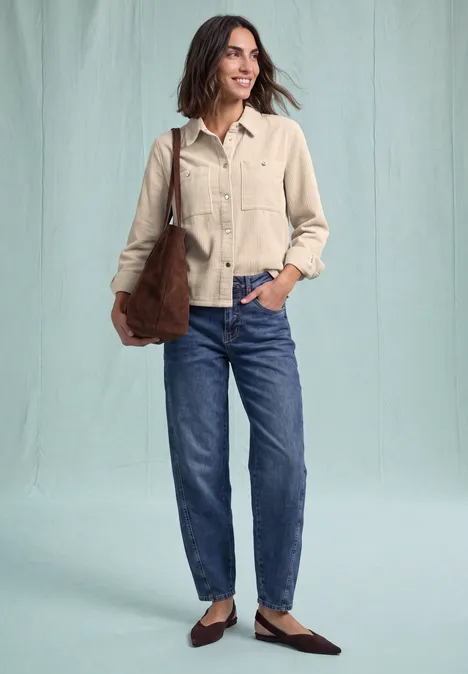 Eine lächelnde Frau in beiger Jacke, blauer Jeans, braunen Schuhen und Schultertasche.