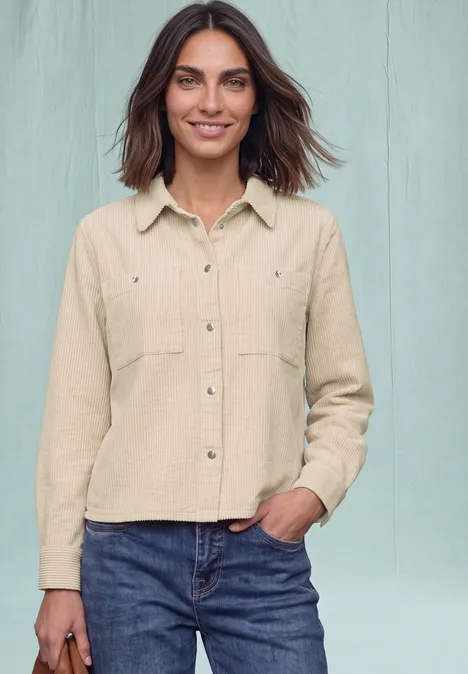 Eine junge Frau trägt eine beige Hemdjacke und blaue Jeans, steht vor hellem Hintergrund und lächelt.