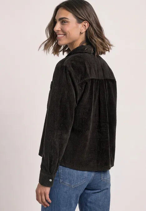 Eine lächelnde Frau in seitlicher Ansicht trägt eine braune Cordjacke und blaue Jeans.