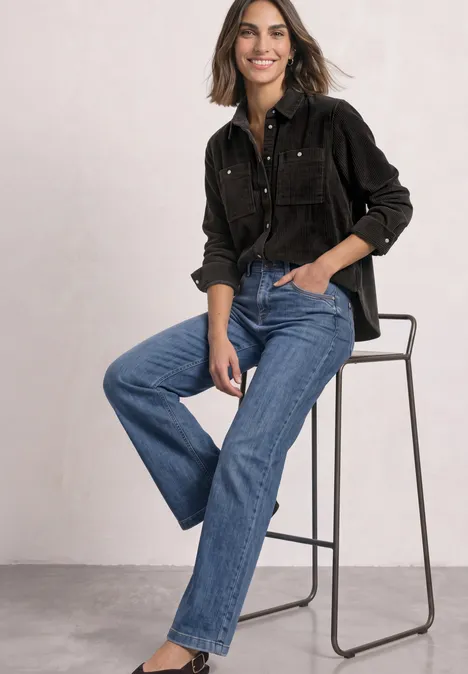 Frau auf hohem Stuhl in schwarzer Bluse und blauen Jeans, sie sitzt entspannt und lächelt.