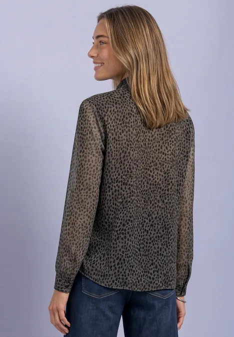 Une femme de profil porte une blouse à motifs discrets, manches longues, et un jean sur fond gris.