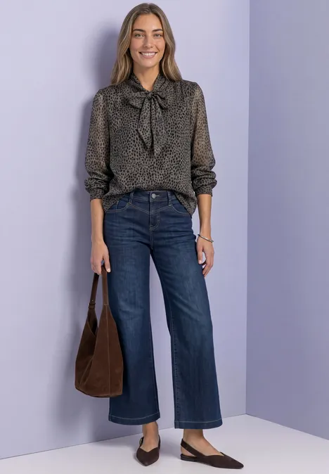 Femme en chemisier à fleurs avec nœud, jean large, sac marron et mocassins pointus sur fond violet.