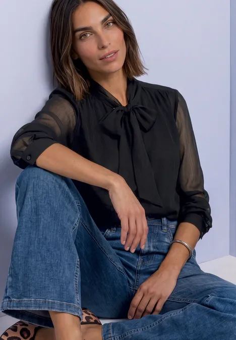 Une femme en chemisier noir à nœud et jean bleu, assise devant un mur clair.
