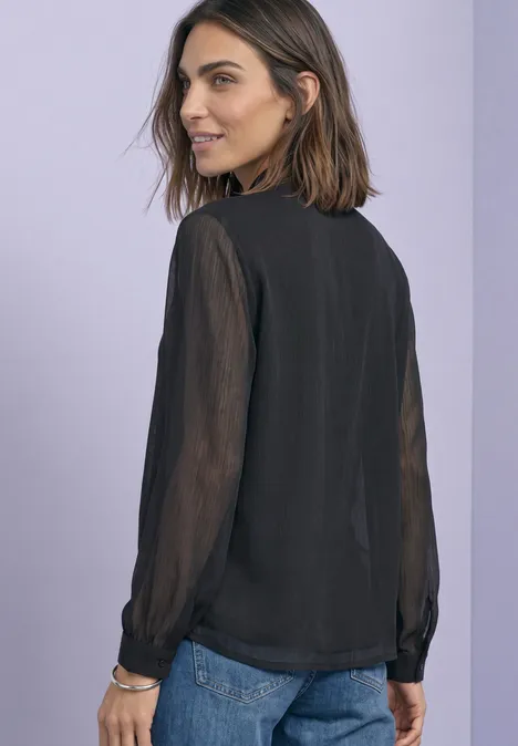 Femme en chemisier noir à manches longues en chiffon, regard latéral sur fond violet clair.