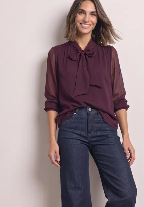 Femme en chemisier bordeaux à col noué et manches longues, associée à un jean bleu foncé sur fond clair.