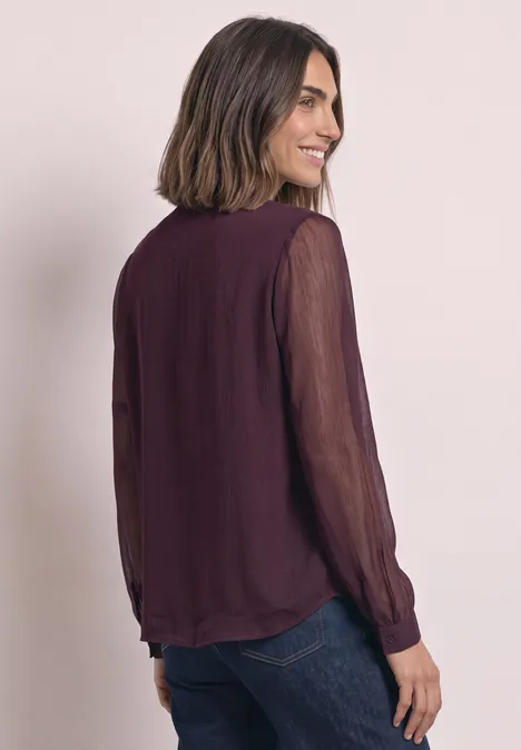 Femme de profil, souriante, portant un chemisier transparent violet foncé à manches longues et un jean sombre.