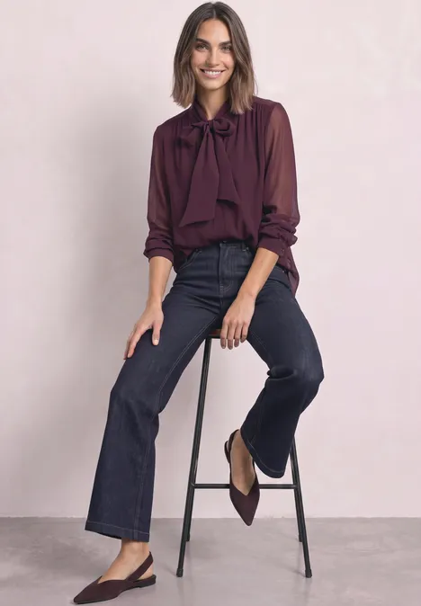Femme assise sur un tabouret, chemisier bordeaux à nouer, jean denim et escarpins slingbacks.