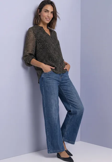 Femme portant un chemisier noir à motifs et un jean large bleu, posant de façon détendue.