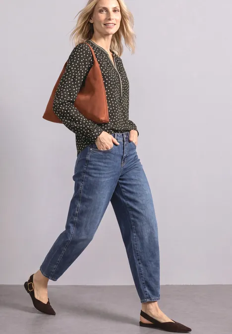 Langarm Tunika mit Split Neck und Print black coffee Frau in schwarzer gepunkteter Bluse, blauen Jeans, schwarzen Ballerinas und brauner Schultertasche vor einfarbiger Wand.