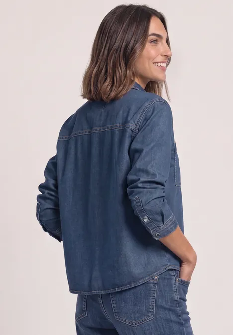 Eine Frau im dunkelblauen Denim-Set (Hemd & Hose), die seitlich lächelnd mit Hand in der Hosentasche steht.