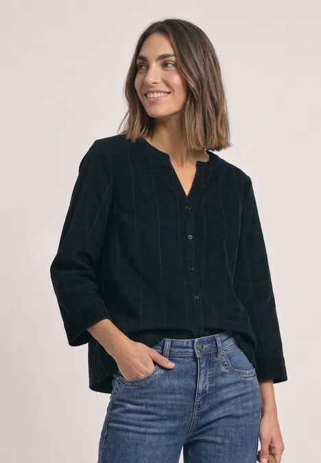 Femme souriante vêtue d’un chemisier noir et d’un jean bleu, debout, mains dans les poches.