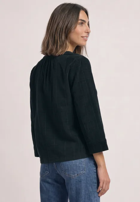 Femme de dos légèrement de profil, portant un chemisier noir texturé et un jean bleu, regard par-dessus l'épaule.