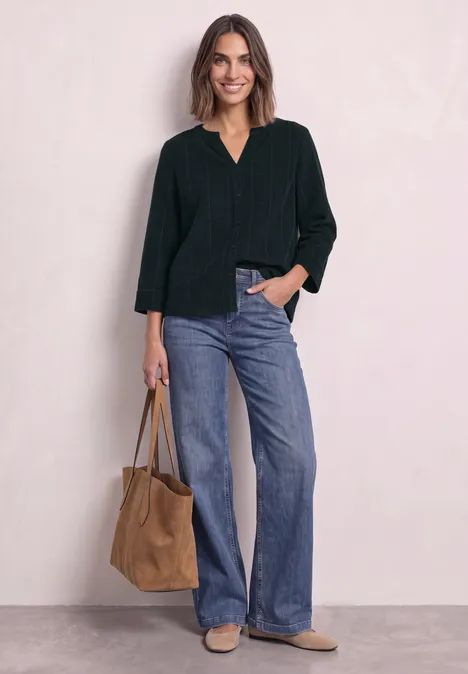 Femme en haut vert foncé et jean bleu, souriante, tenant un sac marron.