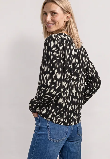 Eine blonde Frau schaut lächelnd über die Schulter, trägt eine schwarz-weiß gemusterte Bluse und Jeans.