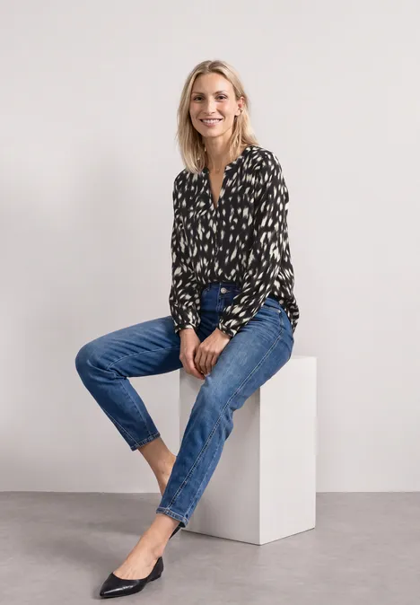 Frau in schwarzer gemusterter Bluse und blauen Jeans sitzt elegant auf weißem Würfel.