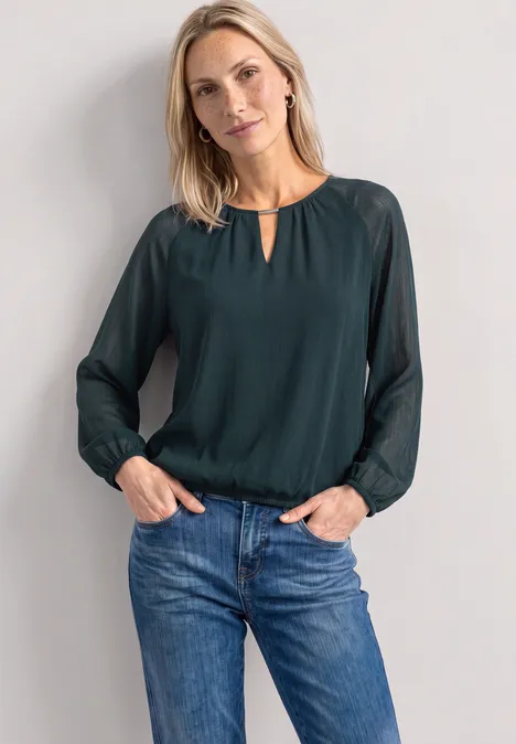 Frau in grüner Bluse und Jeans vor neutralem Hintergrund, Hände in den Taschen.
