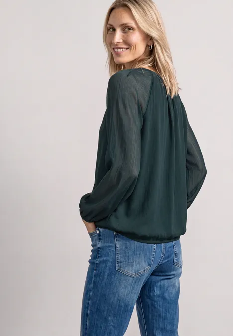 Eine lächelnde Frau in Jeans und dunkelgrüner Bluse blickt über die Schulter in die Kamera.