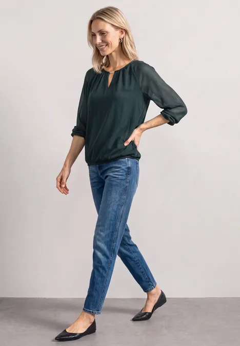 Frau in grüner Bluse und blauen Jeans steht lächelnd vor weißem Hintergrund.