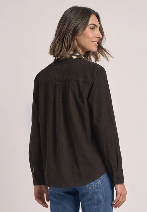 Chemisier à manches longues avec des détails imprimés représentant des chevreuils black coffee Une femme porte une veste-chemise en velours côtelé brun foncé et un jean, elle regarde par-dessus son épaule.
