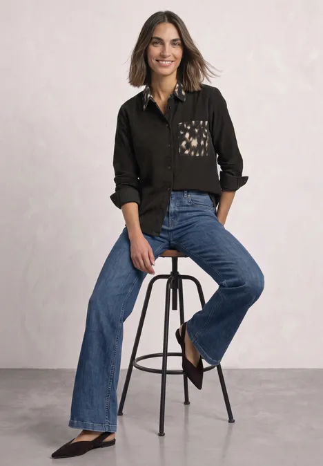 Chemisier à manches longues avec des détails imprimés représentant des chevreuils black coffee Femme en chemise noire à poche imprimée, assise sur un tabouret, portant un jean évasé et souriante.