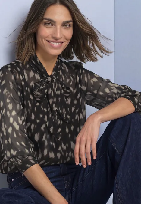 Une femme souriante en chemisier à motifs et jean, posant détendue contre un mur.